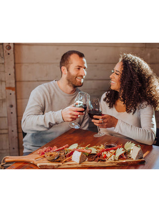 A man and woman toasting with wine glasses, with a wooden plank covered with appetizers in the foreground.