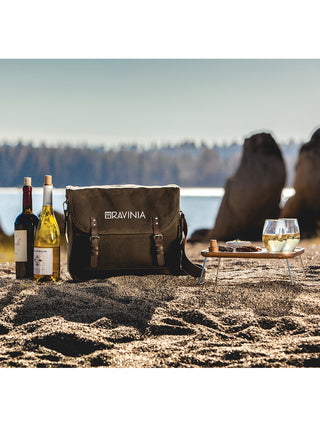 A beach scene featuring a green waxed cotton tote with RAVINIA printed on it in white, two wine bottles next to it, and a small wood table with two wine glasses on top.