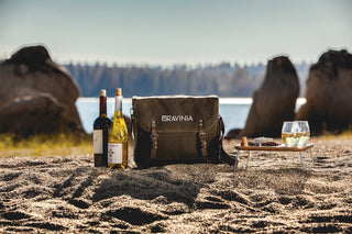 A beach scene with two bottles of wine, a canvas wine tote with the word Ravinia on it, and a small table with two glasses of wine on it.