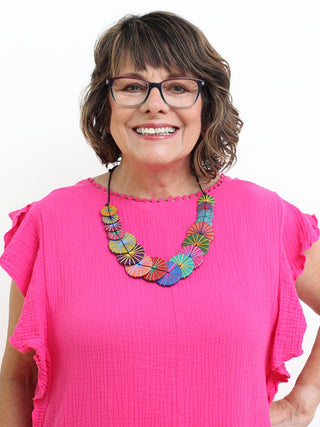A necklace of Round multicolored wooden beads that are layered together and complemented by multiple thread colors that wraps around each bead, on a smiling model in a pink top.