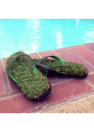 A pair of flip flops that appear to be made of grass, with green and black straps, on bricks next to a pool. 