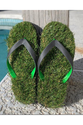 A pair of flip flops that appear to be made of grass, with green and black straps, leaning against a wood board next to a pool.