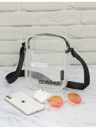 A transparent brick bag with a black strap and the Ravinia logo and name in black. A cell phone and sunglasses are in front of the bag.