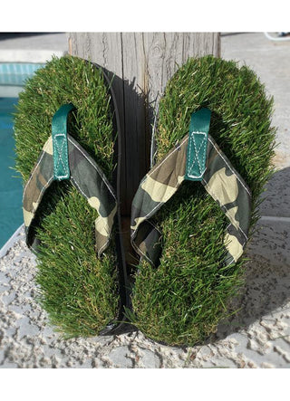 A pair of flip flops that appear to be made of grass, with camoflaged straps, standing up against a board next to a pool.