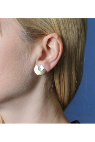 In a blonde-haired model's ear, A dished silver disc on top of a lightly hammered brass semi circle clip earring. 
