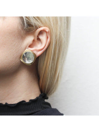 A brass rounded square layered earring with a dished silver-toned disc, on a model with blond hair. 