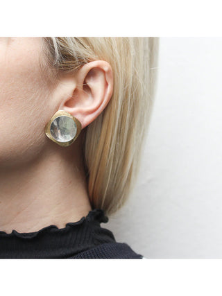 A brass rounded square layered earring with a dished silver-toned disc, on a model with blond hair.