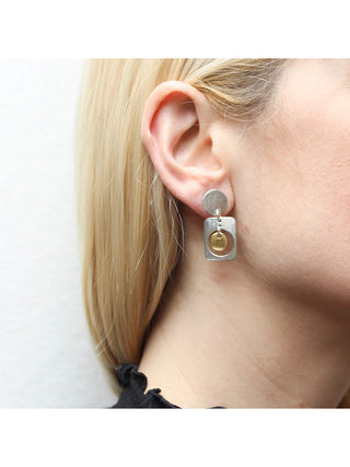 An Earring with a silver-toned disc linked with a cutout rounded rectangle with a brass disc hanging in the center., hanging from a blond-haired model's ear.