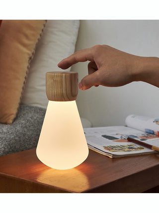 An illuminate light bulb with a wood base, upside down, on top of a desk with a person's hand about to touch it.