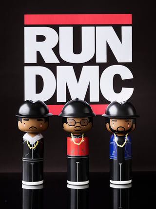 Three wooden dolls representing the group RUN DMC. all with gold chains, against a black background.