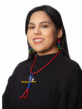 A smiling woman in a black top wearing A necklace with a red chain and a little man pendant comprised of blue, yellow and red, with a clear head. She's also wearing little man earrings.