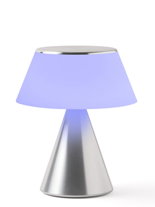 A lamp with a triangular aluminum base and an illuminated  lilac shade.