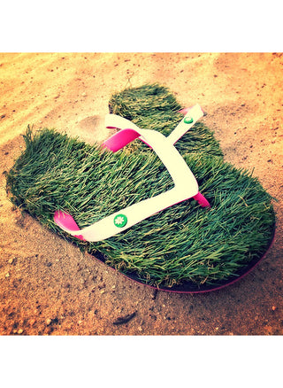 A pair of flip flops that appear to be made of grass, with pinks and white straps, on brown sand.