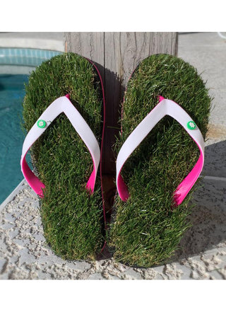 A pair of flip flops that appear to be made of grass, with pinks and white straps, standing leaning against a wood board next to a swimming pool.