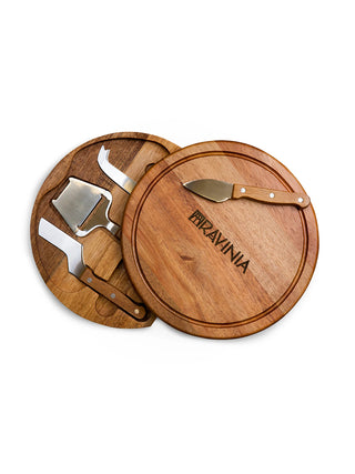A circular acacia wooden cheese board with the Ravinia branding, half-swiveled open to reveal the storage space for four cheese tools.
