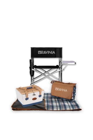 A black chair with a side table, a canvas and wicker picnic basket, and a plaid ground blanket in front of a white background.