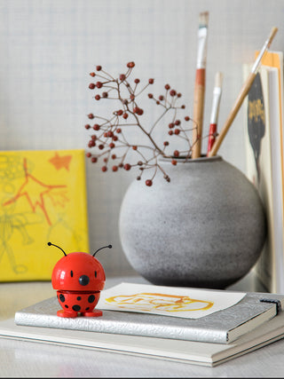 A ladybug-like figure, red with black spots, with a spring in its middle, perched on a shelf in front of a vase.