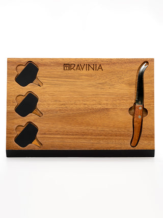 A dark wood rectangular board with the Ravinia logo across the top, a knife in a slot on the right, and three cheese signs on the left, with goat, swiss and brie written on them.