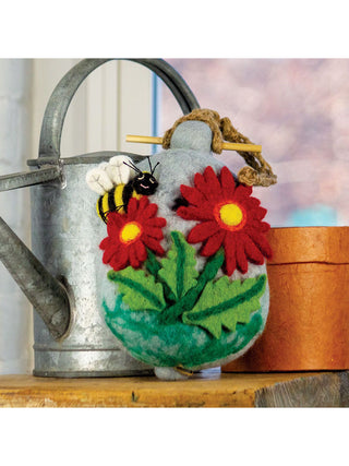 On a shelf in front of a watering can, a felt birdhouse with two red flowers, a leaf underneath and a happy bumblebee.