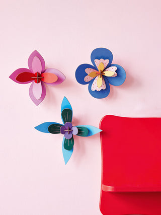 Three colorful paper flowers on a wall, the top two mainly cobalt, and the other red and pink..