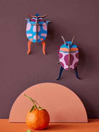 Two cardboard ladybugs, the left primarily red, and the right primarily blue., mounted on a dark red wall with an orange below.