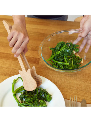A hand scooping greens from a bowl onto a plate with a pair of wood, guitar-shaped tongs.