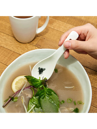 A ceramic spoon with a fly printed on it being dipped into a bowl of vegetable and noodle soup.