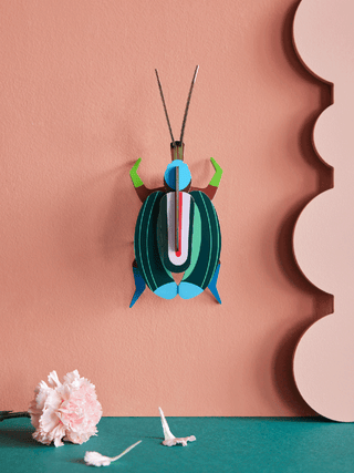 Mounted to an orange wall, a cardboard beetle with green vertical stripes, green front legs and blue back legs.