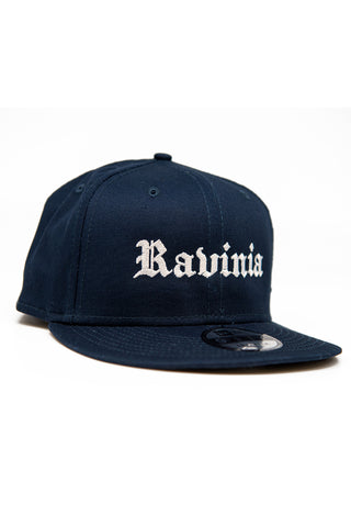 A navy blue baseball cap with Ravinia in white in Gothic script, and a New Era sticker on the bill.