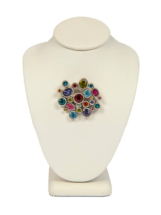 An oval silver brooch with multicolored crystals on a white form.