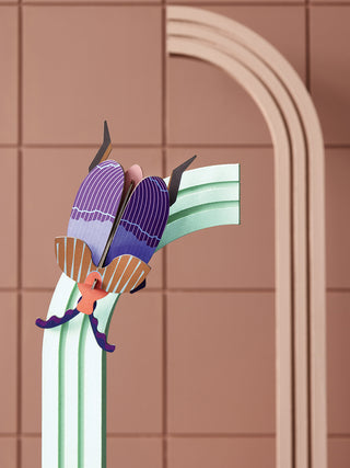 A purple, brown and orange cardboard queen beetle, propped on a wall.