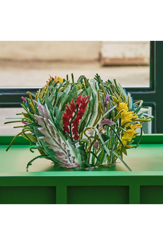 On a green shelf is a vase that looks like it's made by nature, with red, yellow and purple flowers popping up among what appear to be green grasses and plants.