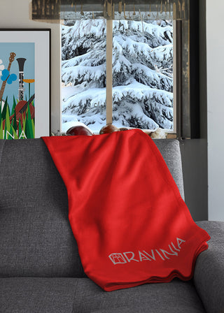 A red blanket with the word Ravinia and the Ravinia logo in gray no it, thrown on a gray sofa, with a snow-covered fir tree visible from the window behind the sofa.