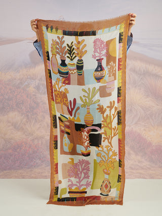 A person holding up a colorful scarf with a tan border and colorful floral details, embroidered, to show the full pattern.
