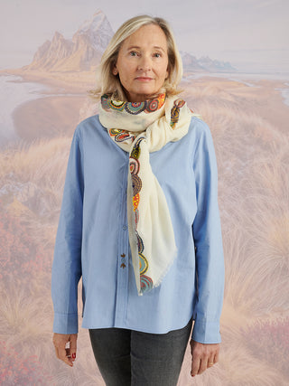 A woman wearing a blue shirt and a natural-colored scarf with colorful embroidered circular details on the border tied around her neck.