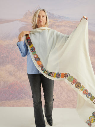A woman holding a natural-colored scarf with a colorful embroidered border to show the full pattern.