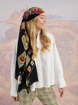 A woman wearing a black scarf with colorful geometric designs and embroidery tied around her head.