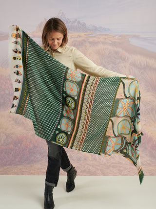 A woman holding up a teal wool scarf to show the tan, black, and blue printed and embroidered patterns.