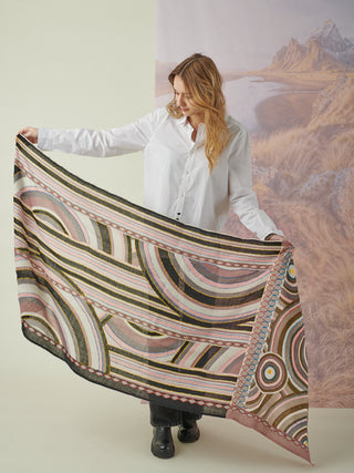 A woman holding a pink, gray, white, and black scarf with geometric details and colorful embroidery, held to show the full pattern.