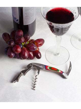 An unfolded wine opener with vibrant colors reminiscent of Marrakesh, below grapes, a bottle of wine and a glass of wine.