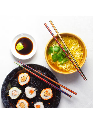 Two pair of colorful wooden chopsticks, one sitting atop a small bowl of noodles, and the other pair atop a large plate of sushi.