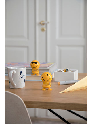 Two smiling yellow figures with round heads, flat feet, and a spring in the middle., on  a table adjacent to a coffee cup