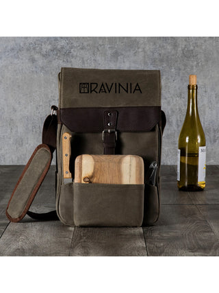 A Ravinia branded bag with a cutting board, a cheese knife, and a corkscrew, in front of a wine bottle, on a gray background.