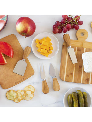 Four cheese tools with wooden handles on and near cutting boards, surrounded by other food like fruit, crackers and pickles.