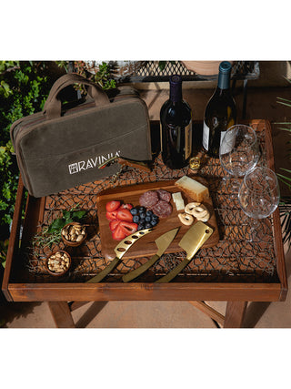 A green waxed cotton bag with RAVINIA on it in white, on a table, surrounded by wine bottles, glasses, and a cutting board filled with appetizers and cheese knives adjacent.