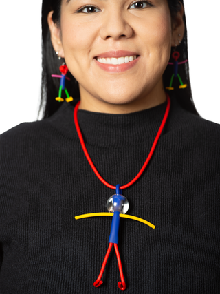 A smiling woman in a black top wearing A necklace with a red chain and a little man pendant comprised of blue, yellow and red, with a clear head. She's also wearing little man earrings.