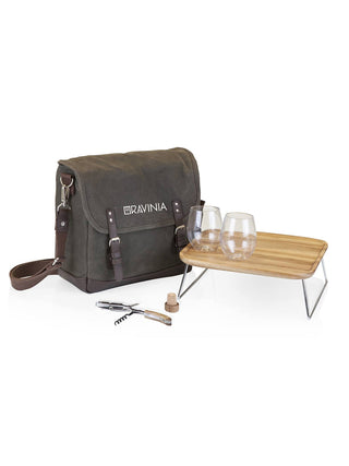 A green waxed cotton tote with RAVINIA printed on it in white, a corkscrew in front of it, and a small wood table with two wine glasses on top.