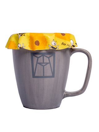 A circular coaster illustrated with goofy, happy looking bees on top of a grey Ravinia mug.