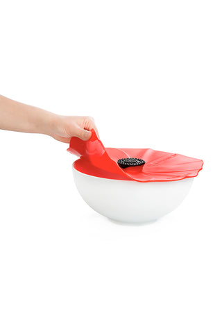 A hand pulling a red poppy silicone lid from a white bowl.