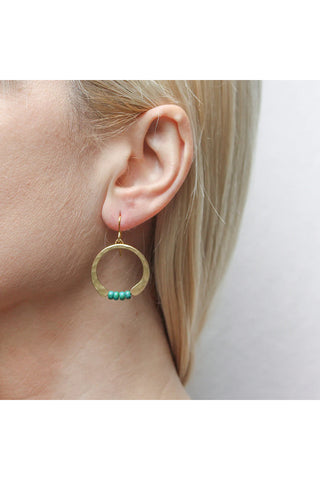 A brass hammered crescent earring completed by four turquoise beads at the bottom, hanging from a blonde-haired woman's ear.
